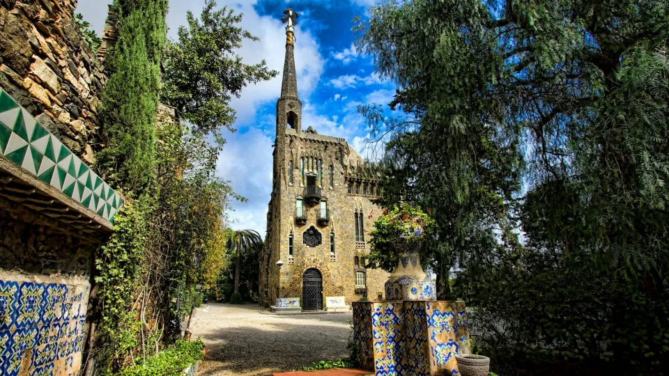 Torre Bellesguard vista desde el camino del jardín, con su torre coronada por la cruz de cuatro brazos y los bancos de trencadís en primer plano